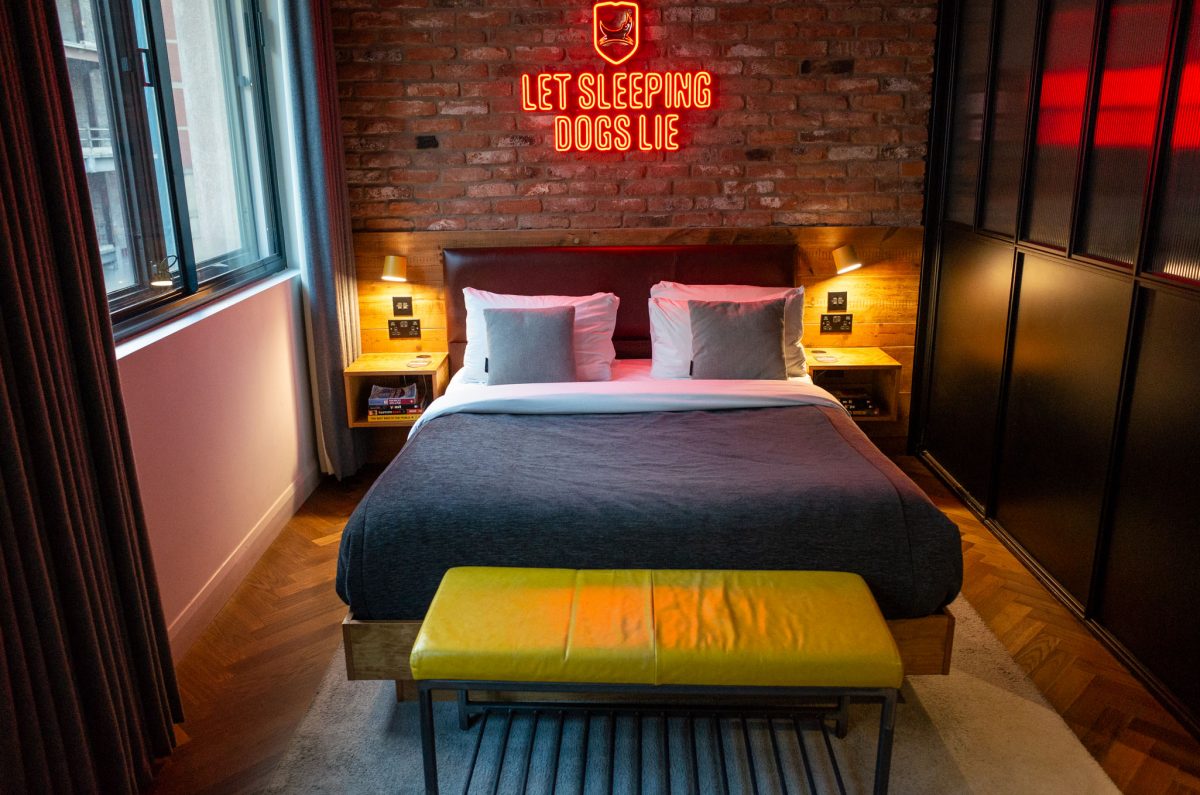 A hotel room with a large bed, brick wall, and a red neon sign above the headboard.