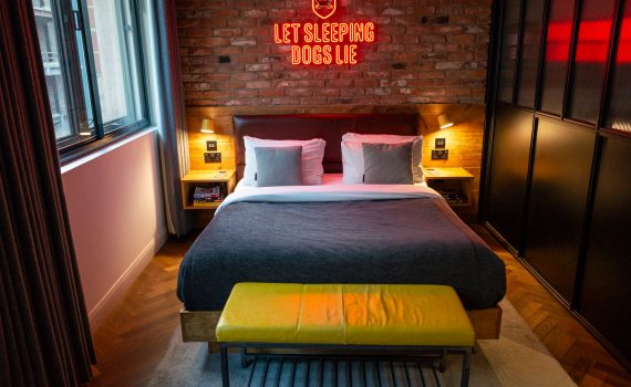 A hotel room with a large bed, brick wall, and a red neon sign above the headboard.