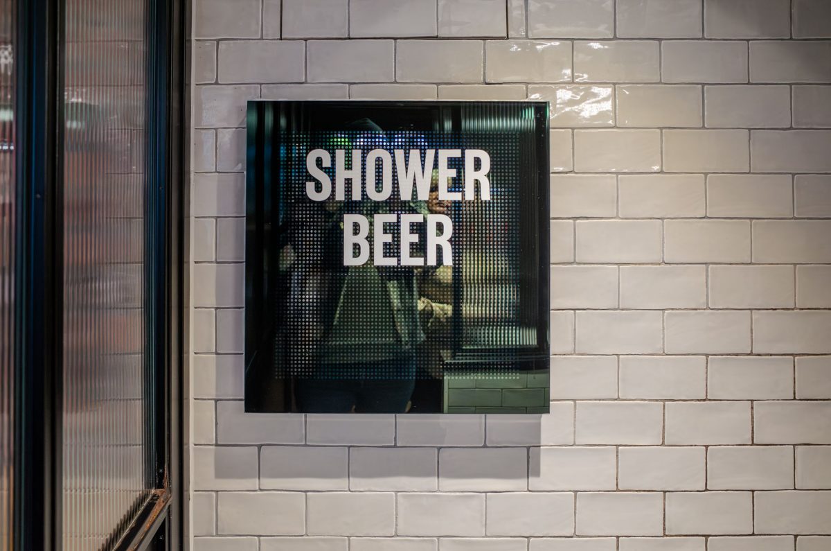 A square sign that says SHOWER BEER mounted on a white subway-tiled wall.