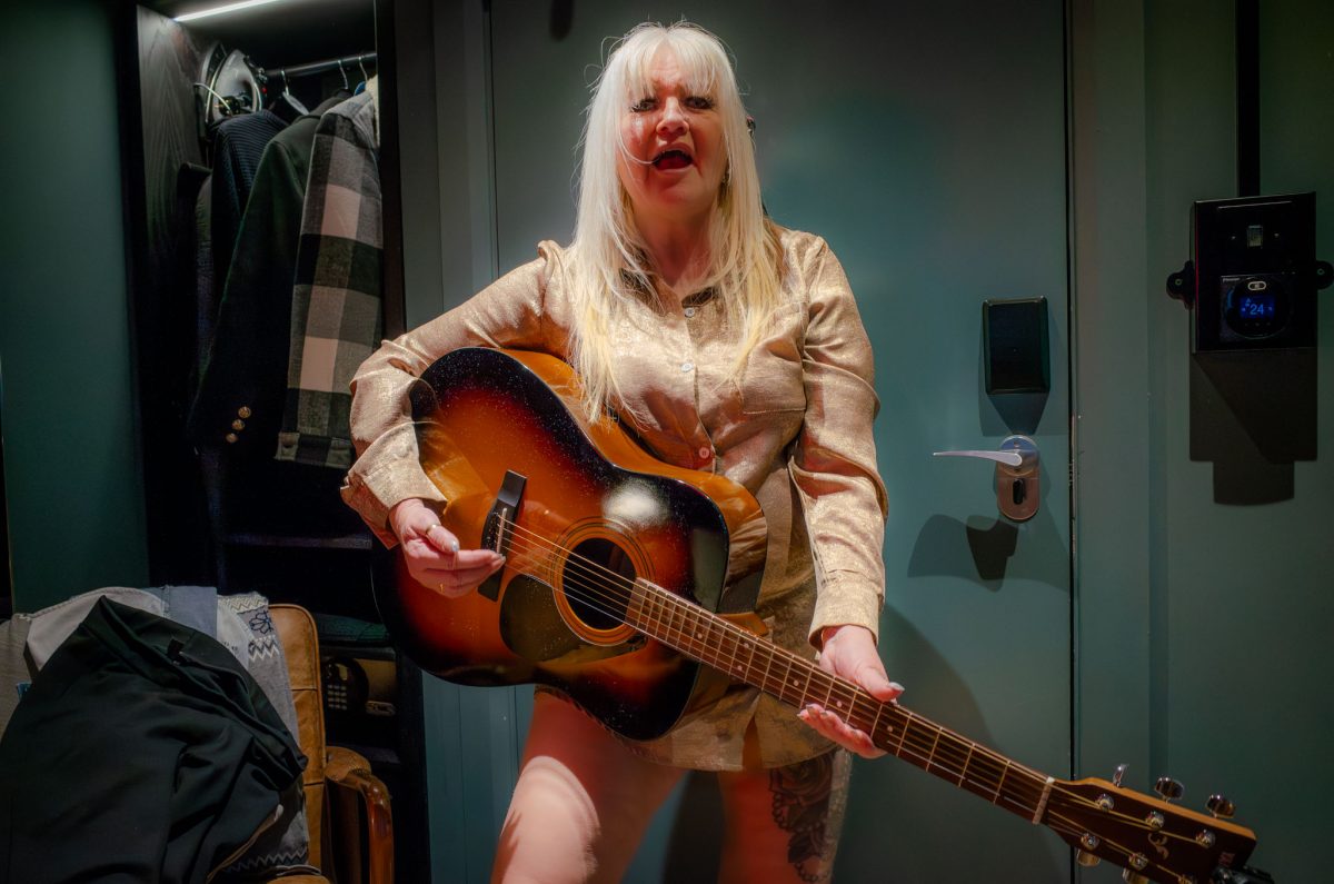 A woman with blonde hair wearing a gold shirt holding an acoustic guitar in a hotel room.