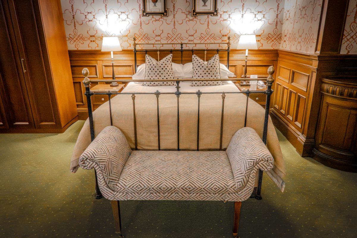 A traditional hotel bedroom with a wrought iron bed, patterned wallpaper, and a decorative bench at the foot of the bed.