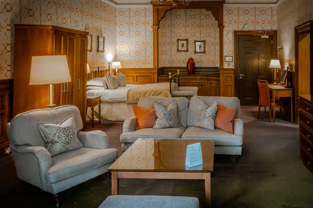 Spacious hotel suite in Edinburgh featuring a plush sofa, armchair, and a bed in the background with Victorian-style wallpaper.