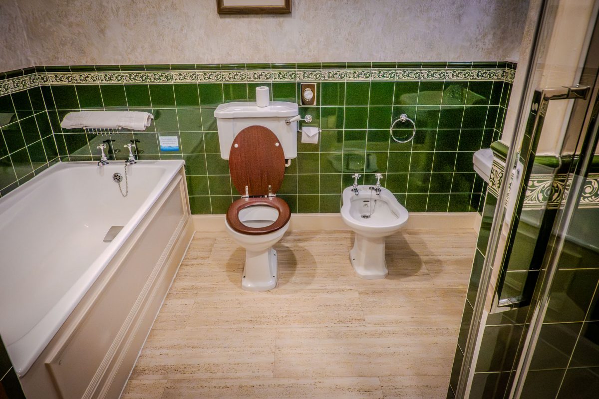 A classic bathroom featuring dark green wall tiles, a white bathtub, toilet, and bidet with vintage-style fixtures.