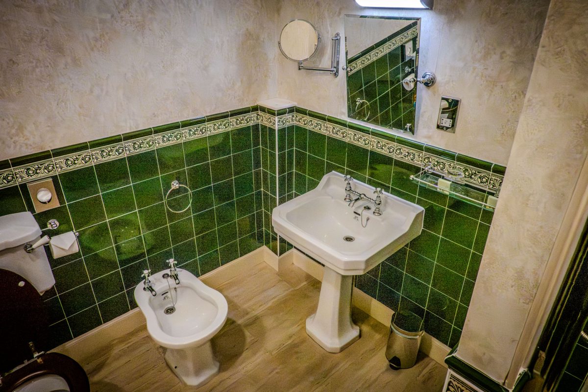 A bathroom corner showing a pedestal wash basin, bidet, and green wall tiling with a chrome mirror.