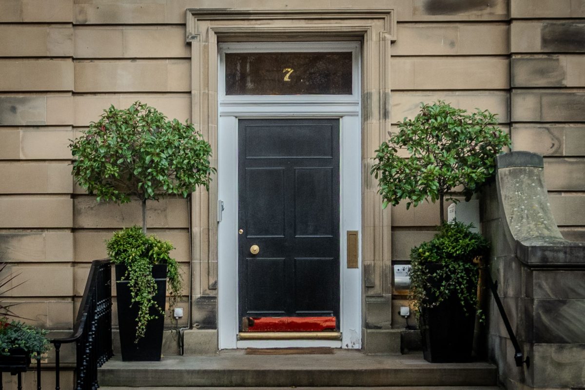 A black front door of a stone townhouse in Edinburgh, numbered 7, flanked by two manicured green plants in black pots.