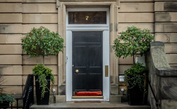 A black front door of a stone townhouse in Edinburgh, numbered 7, flanked by two manicured green plants in black pots.