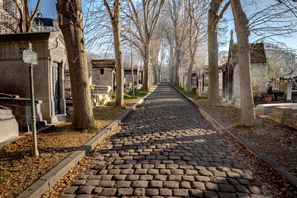 A long perspective view of a cobblestone path lined with bare trees and stone mausoleums at Père Lachaise Cemetery in Paris.