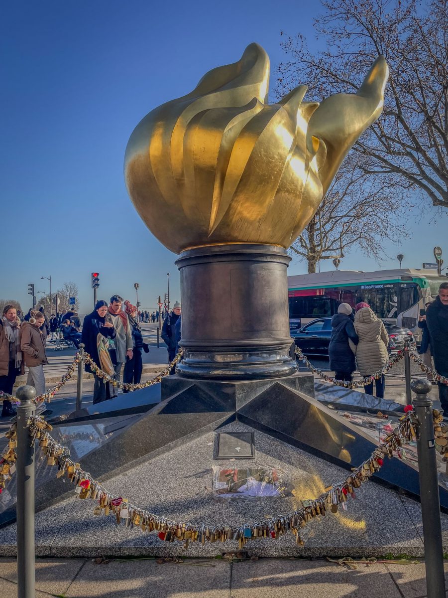 The golden Flame of Liberty monument in Paris surrounded by tribute locks and visitors.