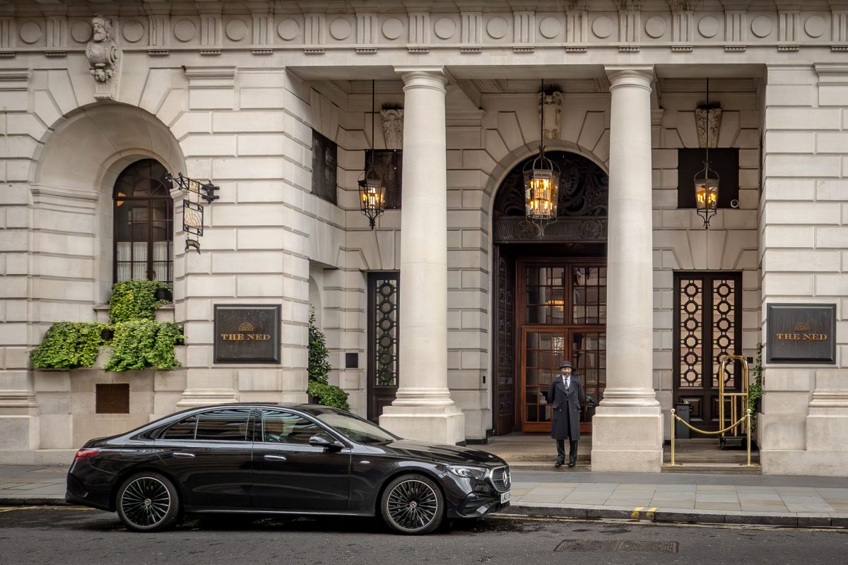 The exterior of The Ned London featuring white stone columns, a doorman, and a luxury black car.