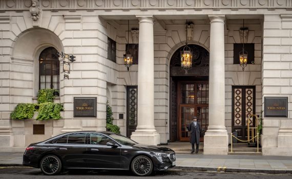 The exterior of The Ned London featuring white stone columns, a doorman, and a luxury black car.