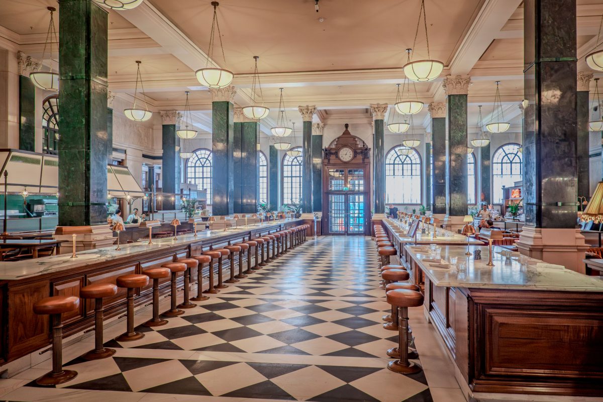 A vast hall with green marble pillars, checkerboard floors, and long dining counters.